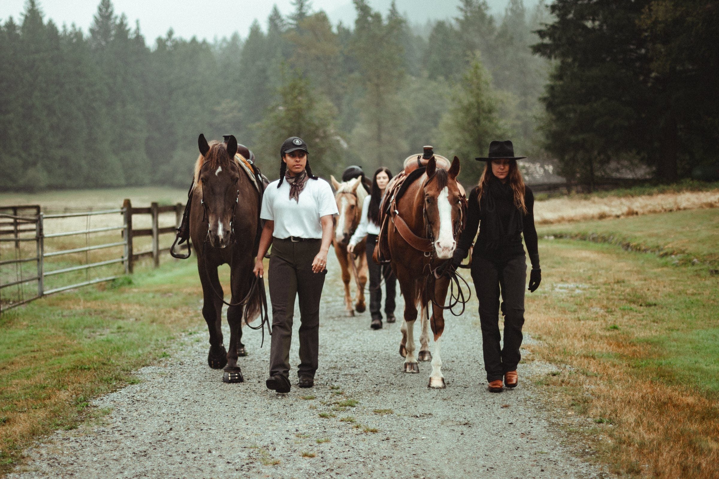 Chelsea and three riders in The Bushwacker lead their horses down a gravel lane past a fence and misty evergreens