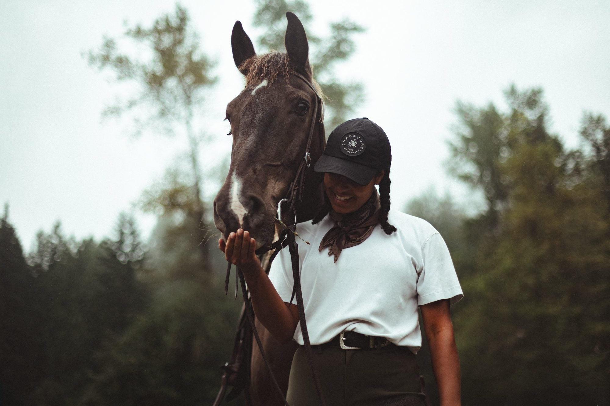 Chelsea, smiling in a Hackmore cap, holds her hand up to her dark bay horse's muzzle